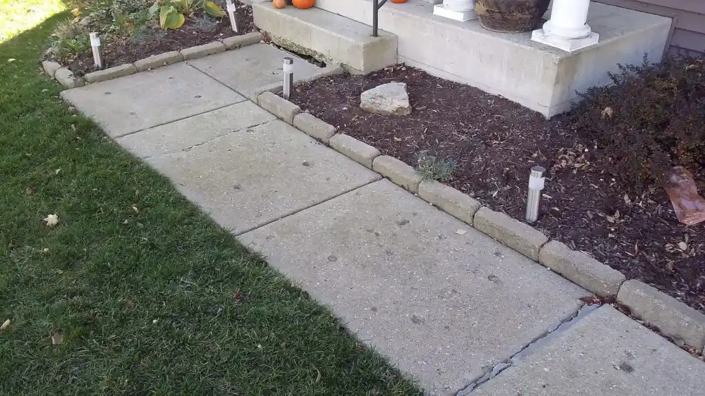 A concrete walkway bordered by stone edging leads to a porch with pumpkins. Grass is on the left, mulch and small plants on the right, and small garden lights line the path—reminding us why mudjacking fails in dynamic landscapes like this.