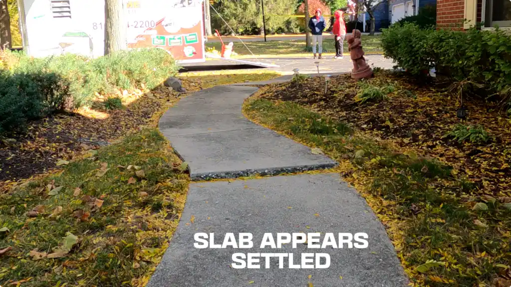 A concrete walkway curves through a yard with autumn leaves scattered on the ground. The text SLAB APPEARS SETTLED is overlaid on the path. Fixing Raised Concrete in McHenry ensures smooth, safe paths bordered by shrubs and trees.