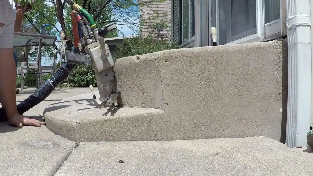 A concrete leveling machine addresses one of the Top 3 Concrete Leveling Problems as it lifts a sunken step outside a house, with hoses and cables attached, while a worker kneels nearby on the left.