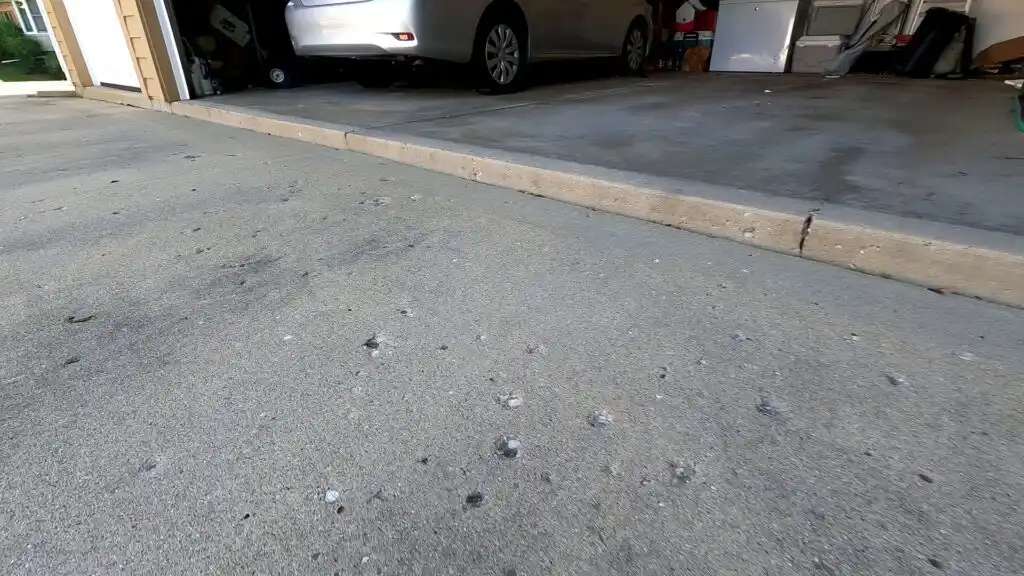 A concrete driveway with dark spots and signs of sunken concrete problems leads to an open garage. A silver car is parked inside, and various items are visible along the garage walls. The sun casts light on part of the driveway.