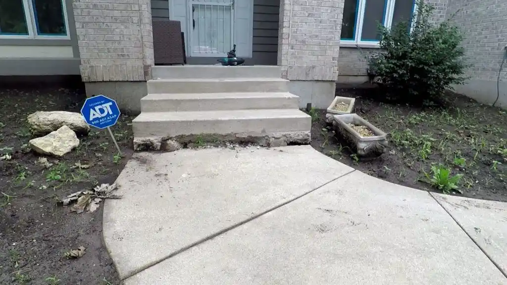 Concrete steps leading to a front door have settled unevenly, creating a noticeable gap beneath the steps—a clear sign of sunken concrete problems. An ADT security sign is visible on the left, with planters and sparse plants around the walkway.