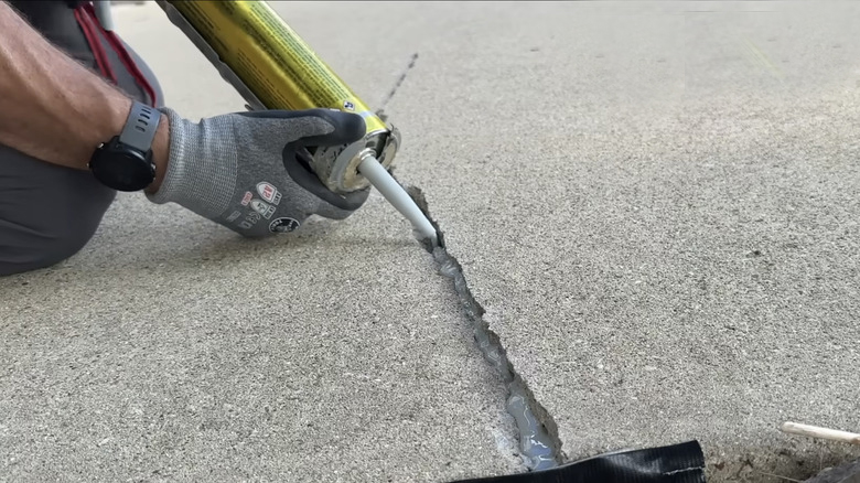 A person wearing a gray glove uses a yellow caulking gun for concrete caulking, applying sealant to a crack in a concrete surface.