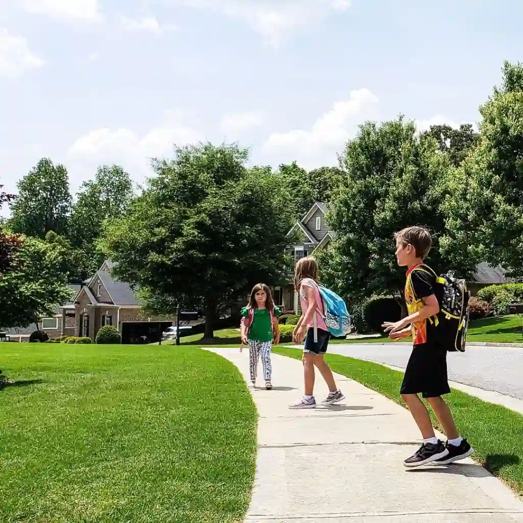 Three children with backpacks walk along a sunny suburban sidewalk bordered by grass and trees, with houses visible in the background.