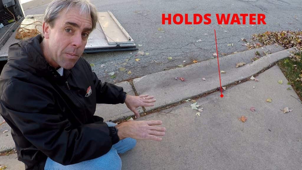 A man crouches by a sidewalk, gesturing to a dip near the curb. Red text and an arrow point to a spot labeled HOLDS WATER. Some fallen leaves and a street are visible in the background.