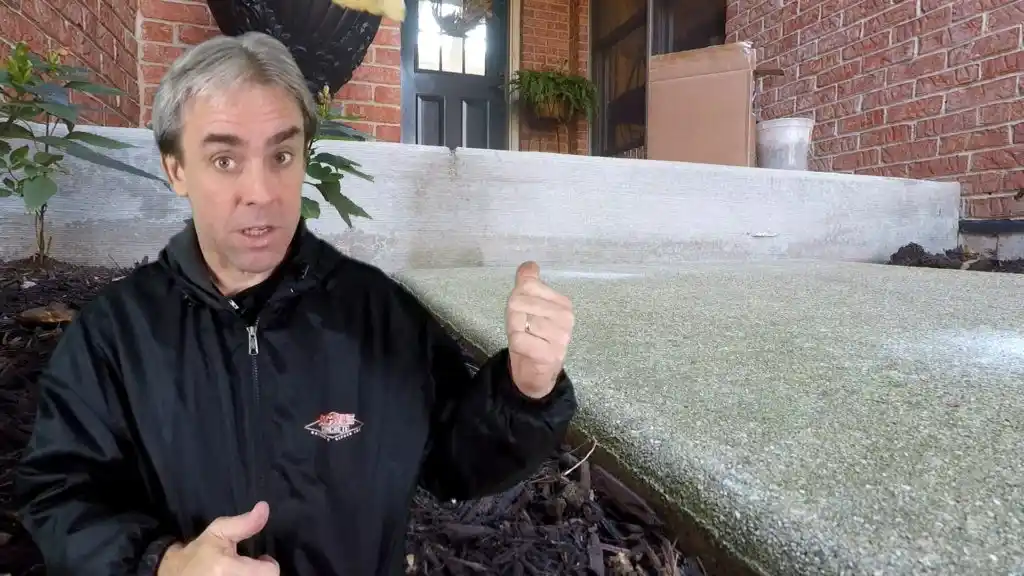 A man in a black jacket gestures toward a sloped concrete porch step in front of a brick house with a black door and cardboard box on the porch.