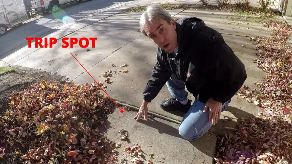 A man kneels on a sidewalk, pointing to a raised section labeled TRIP SPOT in red text with an arrow. Fallen leaves are scattered around, and a street with parked cars is visible in the background.