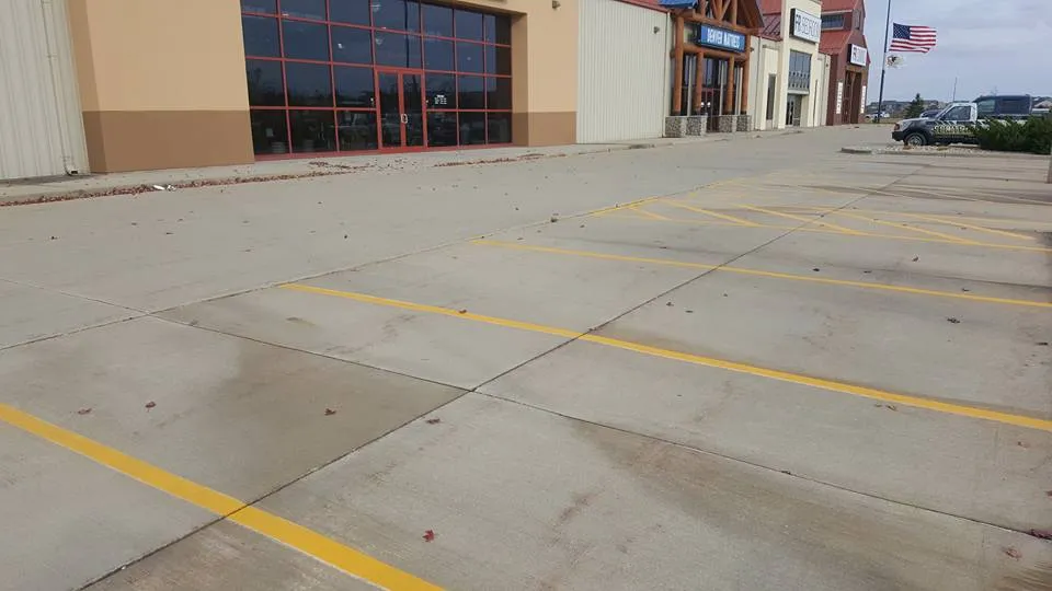 Empty parking lot with freshly painted yellow lines in front of stores. An American flag is flying in the background on the right side, and the sky is clear. The area showcases recent commercial parking lot concrete repair, giving a clean and well-maintained appearance.