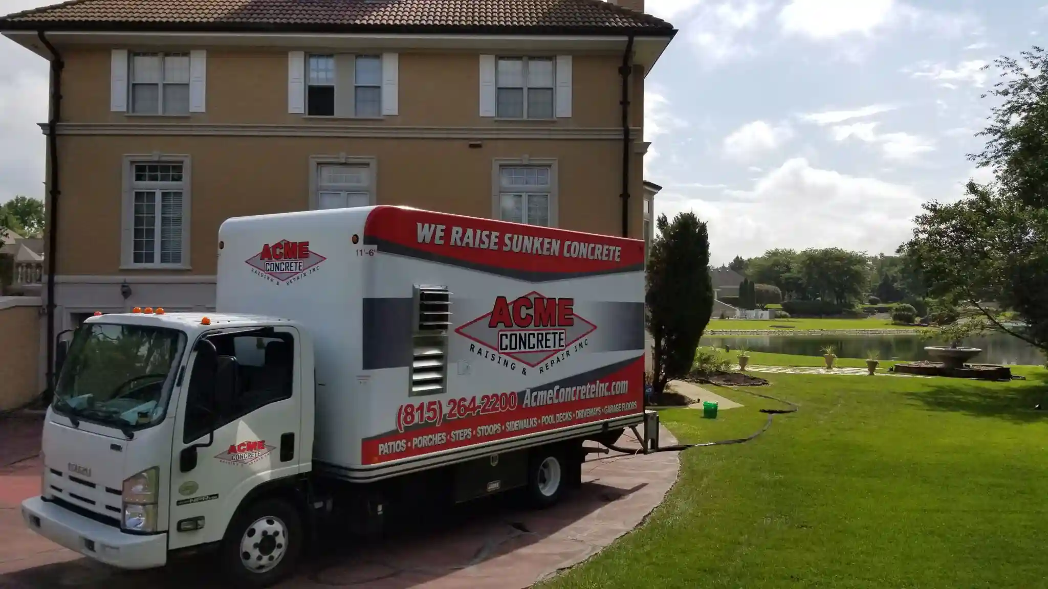 A white ACME Concrete truck with red signage, specializing in raising concrete in South Elgin, is parked in front of a large beige house, with a green lawn, trees, and a pond visible in the background.