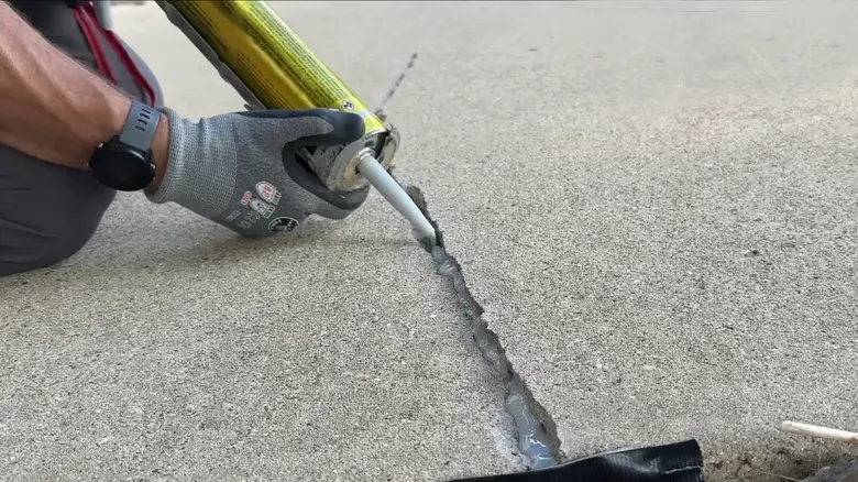 A person wearing gloves is applying sealant from a tube to fill a crack in a concrete surface, demonstrating professional Concrete Caulking in Prairie Grove.