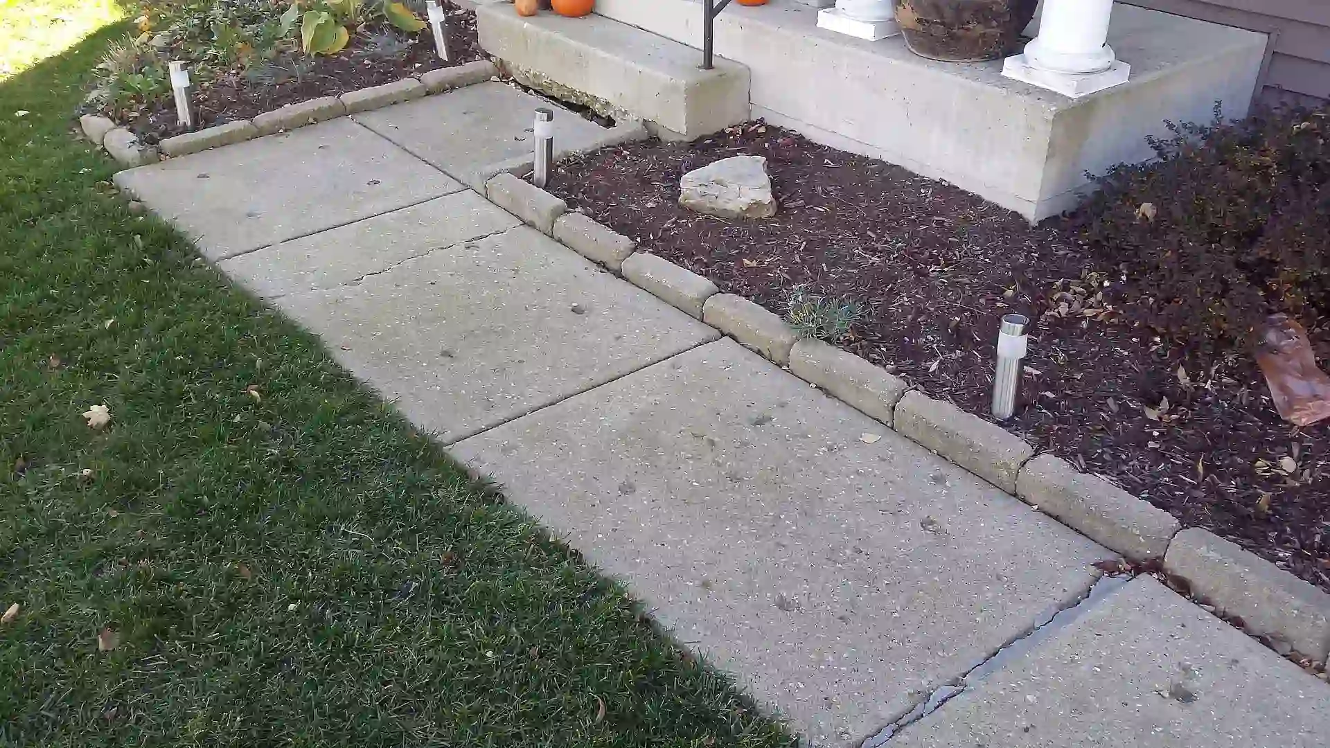 A concrete walkway bordered by grass and mulch leads to a porch with pumpkins and potted plants, showcasing the quality you’d expect from raising concrete in Lincolnshire. Small garden lights line the walkway edge, illuminating a well-maintained entrance.
