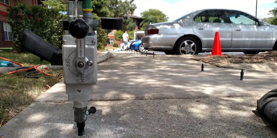 A close-up of a concrete leveling tool inserted into a sidewalk, with cars and orange traffic cones in the background—demonstrating the process of raising concrete in South Barrington.