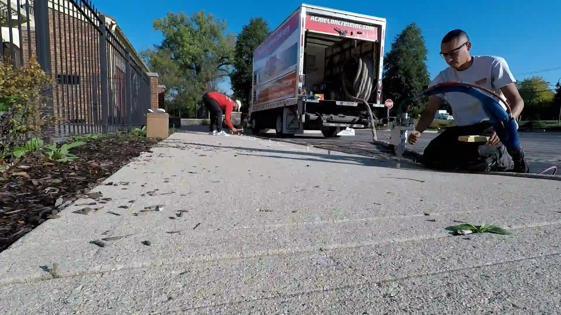 Two people work outside next to a moving truck, raising concrete in Round Lake Heights; one kneels near tools while the other bends over by the truck. A small dog stands on the sidewalk near them. Trees and a fence are visible in the background under a clear sky.