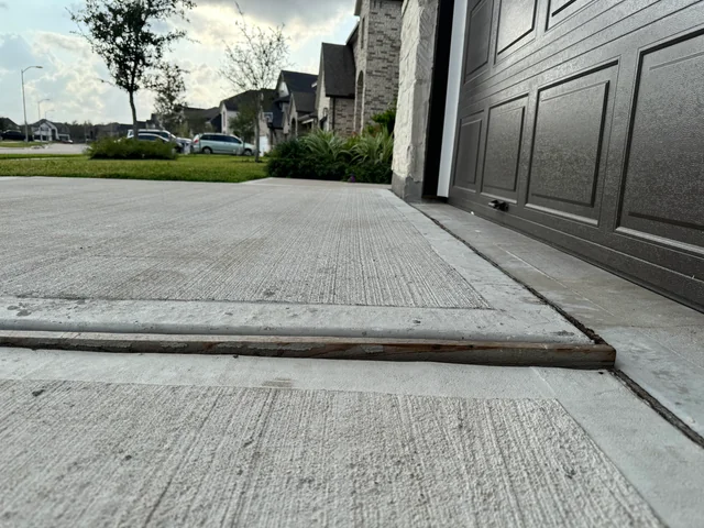 A close-up view of a driveway with a raised concrete slab near a garage door, creating a noticeable tripping hazard due to garage floor lifting. Houses, trees, and cars are visible in the background.