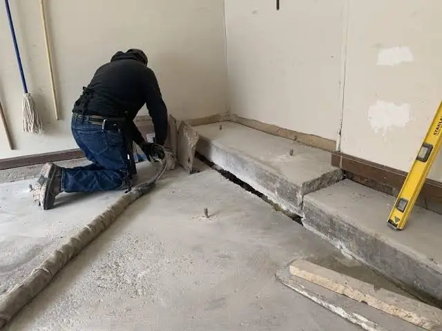 A person in dark clothing kneels on the floor, repairing or cutting a section of cracked concrete near a wall in what appears to be a garage or basement. Tools and construction materials are nearby, suggesting that Garage Floor Lifting in Crystal Lake may be underway to restore stability and ensure a level surface.
