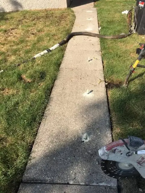 A concrete sidewalk with several small foam patches, a hose, and a construction saw visible, surrounded by grass. The scene suggests multifamily concrete leveling and sidewalk repair work is in progress.