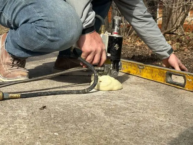 A person kneels on concrete using a yellow leveling tool and equipment, possibly to repair or level the surface, as part of HOA Concrete Services. A hose and some foam material are also visible on the ground.