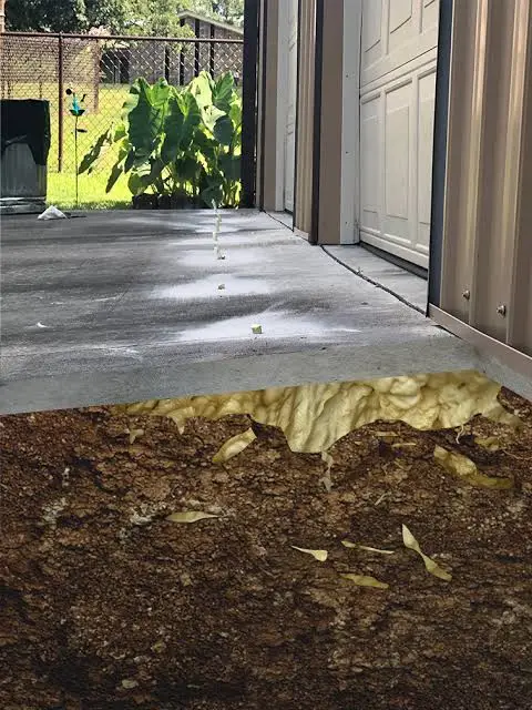 A cross-section of a concrete driveway shows foam insulation beneath the slab and soil underneath—an effective method often used in garage floor lifting—next to a metal garage and a grassy yard with large green plants.